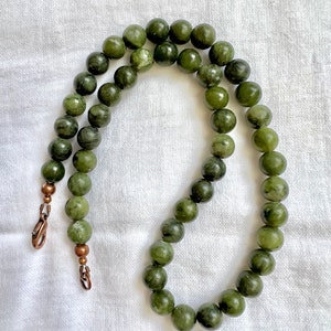 Puede incluir: Un collar de cuentas de jade verde con cierres de cobre. Las cuentas son redondas y tienen un color verde moteado.