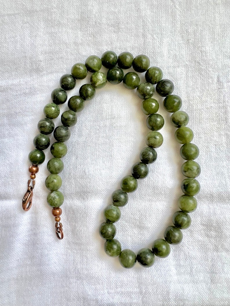 Puede incluir: Un collar de cuentas de jade verde con cierres de cobre. Las cuentas son redondas y tienen un color verde moteado.