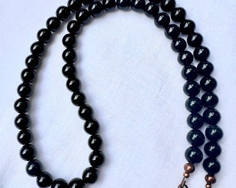 Puede incluir: Un collar de cuentas negras con un cierre color cobre. El collar está compuesto por múltiples hilos de cuentas negras redondas y brillantes. Las cuentas son de tamaño uniforme y están ensartadas, creando un accesorio elegante.