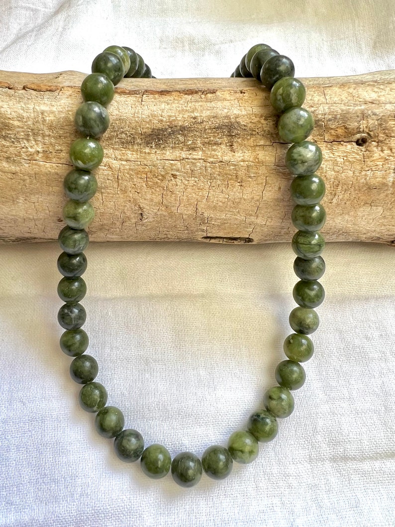 Puede incluir: Un collar de jade verde con cuentas redondas. El collar est&aacute; drapeado sobre un trozo de madera a la deriva.