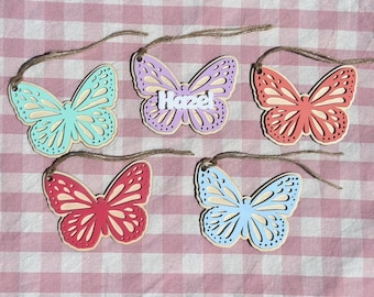 May include: Five wooden butterfly ornaments with different colored paint. The butterflies are hanging from twine. One butterfly has the name "Hazel" written on it.