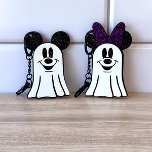 May include: Two black and white ghost keychains with Mickey Mouse ears. One keychain has a purple glitter bow.