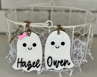 May include: Two white wooden ghost-shaped tags with black eyes and smiles, personalised with the names "Hazel" and "Owen" in black script. One ghost has a pink bow. They hang from a white wire basket with paper filler.