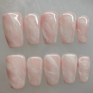 May include: A set of ten artificial nails in a light pink hue with a marbled white design. The nails are rectangular with rounded edges and a glossy finish. The set includes various sizes for a custom fit.