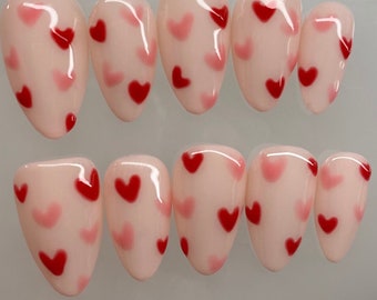 May include: A set of ten artificial nails with a glossy finish. Each nail is a pale pink color and decorated with a pattern of red and pink hearts. The nails are oval-shaped.