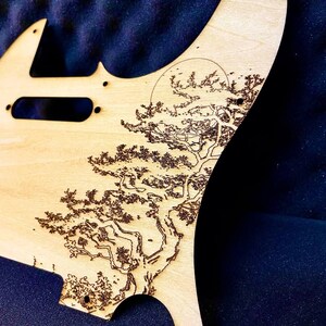 Può includere: Un battipenna per chitarra in legno chiaro con un design dettagliato inciso al laser di un albero e un sole. L'opera d'arte è di colore marrone scuro, creando un contrasto con la venatura naturale del legno. Il battipenna ha diversi fori di montaggio.
