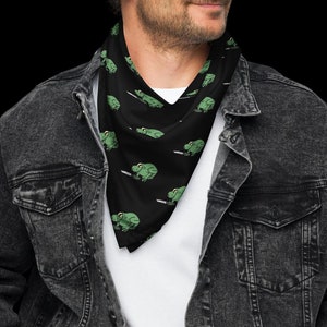 May include: Black bandana featuring a repeating pattern of green cartoon frogs with knives. The bandana is worn around the neck. The person in the image is wearing a white t-shirt and a denim jacket.