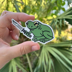 May include: A green frog sticker holding a black knife. The frog has a toothy grin and large, yellow eyes.