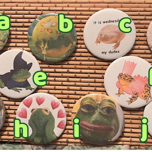 May include: Ten round buttons with different images of frogs and other animals. The buttons are arranged in a grid pattern on a brown woven surface. The buttons are labeled with letters a through j.