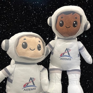May include: Two plush astronaut dolls in white space suits with gray accents. The dolls have smiling faces and wear helmets. The Artemis logo is printed on the chest of each doll. The background is a starry night sky.