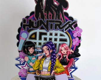 May include: A colorful cake topper featuring three anime-style women holding microphones. The topper has the word "HUNTX" and the name "VERA". The design includes pink flowers, stars, and a silhouette of a band.