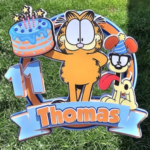 May include: A blue and orange birthday cake topper featuring Garfield the cat and Odie the dog. The cake topper has a blue and orange cake with candles, the number 11, and the name Thomas.