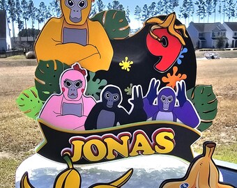 May include: A cake topper featuring a yellow, pink, black, and purple monkey with a banana peel on top. The topper also includes a black banner with the name "JONAS" and the number "9" with colorful paint splatters.