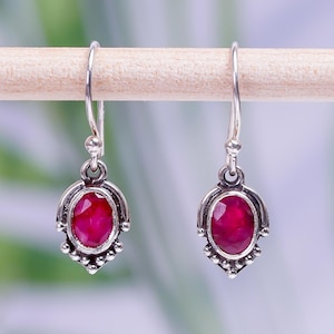 May include: A pair of silver drop earrings with oval, deep red gemstones. The earrings have a decorative, ornate design around the gemstones and are suspended from simple hooks. The background is a soft, blurred mix of green and blue.