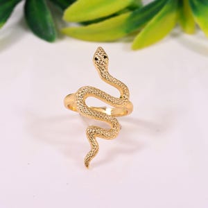 May include: A gold-colored ring in the shape of a snake. The snake's body is textured, and its head is raised with two small black accents for eyes. The ring is set against a white background with green and yellow leaves.