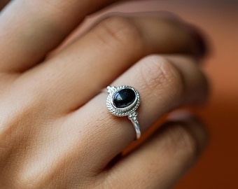 Könnte beinhalten: Ein silberner Ring mit einem ovalen, schwarzen Edelstein. Der Ring hat einen detaillierten, seilartigen Rand um den Stein und dekorative Akzente am Band. Der Ring wird an einem Finger gezeigt.