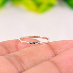 May include: A silver ring with a textured, geometric design. The ring is thin and delicate, with a series of small, elongated shapes forming the band. The ring is held in a hand, with a blurred background of green and white.