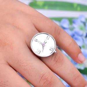 Könnte beinhalten: Ein silberner Ring mit einem Zifferblatt-Design. Der Ring hat ein rundes Zifferblatt mit römischen Ziffern und Uhrzeigern. Ein kleiner, rosa Edelstein ist in der Mitte des Zifferblatts eingesetzt.
