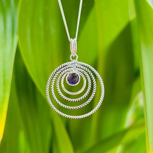 Może przedstawiać: Srebrny naszyjnik z zawieszką przedstawiającą fioletowy kamień szlachetny otoczony trzema koncentrycznymi okręgami.