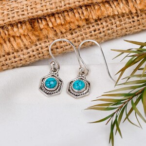 May include: A pair of silver earrings with turquoise-colored, hexagonal stones. The stones are set within a silver frame with a twisted rope design. The earrings have a simple hook closure and are displayed on a white surface.