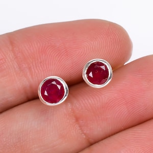 May include: A pair of round stud earrings with faceted, deep red gemstones set in a silver bezel. The earrings are shown against a neutral background, highlighting the vibrant colour and design of the jewellery.