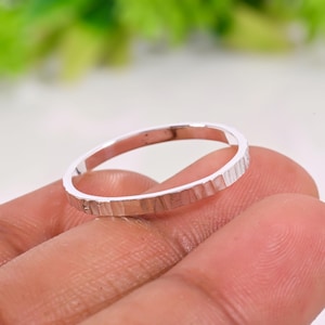 May include: A slim, silver ring with a textured finish. The ring is displayed on a person's finger. The design features a series of vertical lines, giving it a unique, handcrafted look. The ring is a simple band, suitable for everyday wear.