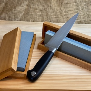 May include: A wooden box containing a grey sharpening stone and a silver knife with a black handle. The knife is resting on the stone.