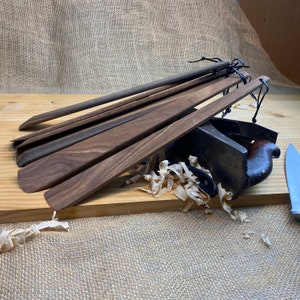 May include: A selection of dark brown wooden spatulas and a plane, arranged on a wooden surface. The tools are tied together with black string. Wood shavings are scattered around the tools. The tools appear to be handcrafted.