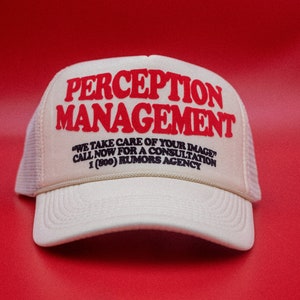 May include: A cream-coloured trucker hat with the words "PERCEPTION MANAGEMENT" in red block letters. Below, the text reads "WE TAKE CARE OF YOUR IMAGE CALL NOW FOR A CONSULTATION 1 (800) RUMORS AGENCY". The hat has a mesh back and a curved brim.