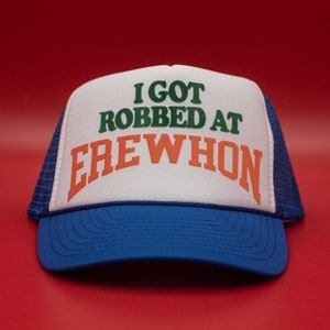 May include: A blue and white trucker hat with the slogan "I GOT ROBBED AT EREWHON" in green and orange. The hat features a blue peak and mesh back, set against a red background.
