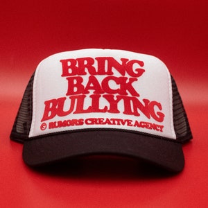 Könnte beinhalten: Ein Trucker-Cap mit weißer Front und schwarzem Mesh-Rücken. Auf der Front steht in roter Schrift "BRING BACK BULLYING" über "RUMORS CREATIVE AGENCY". Der Hut liegt vor rotem Hintergrund.