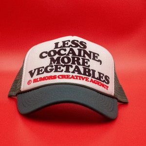 May include: A trucker hat with a white front panel and a dark green brim and mesh back. The hat features the text "LESS COCAINE, MORE VEGETABLES" in black, with "RUMORS CREATIVE AGENCY" in red.