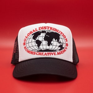 May include: A black and white trucker hat with the text "GLOBAL DISTRIBUTION RUMORS CREATIVE AGENCY" in red. The hat features a world map graphic and a mesh back.