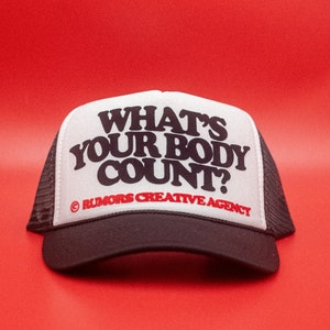 May include: A black and white trucker hat with the text "WHAT'S YOUR BODY COUNT?" in bold, black letters. The hat also features the text "© RUMORS CREATIVE AGENCY" in red. The hat is set against a red background.