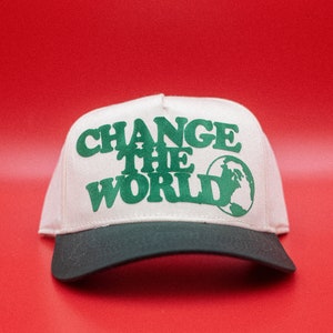 May include: A cream and green baseball cap featuring the text "CHANGE THE WORLD" and a globe design. The cap has a dark green peak and is displayed against a red backdrop.
