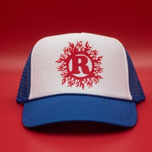 May include: A blue and white trucker hat with a red design. The hat has a white front panel with a red sunburst design around the letter "R". The brim and the lower part of the hat are blue, and the back is a blue mesh.
