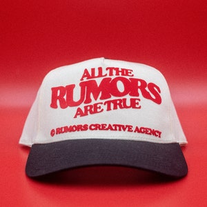 May include: A white and navy blue baseball cap with the text "ALL THE RUMORS ARE TRUE" in red. The cap also features the text "@ RUMORS CREATIVE AGENCY". The hat is set against a red background.