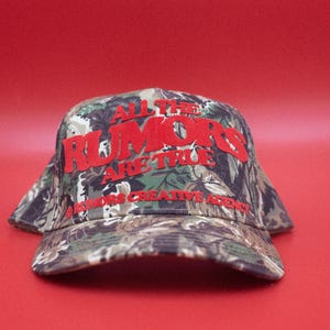 May include: A camouflage baseball cap with the text "ALL THE RUMORS ARE TRUE" in red block letters. The hat also features the text "B. RUMORS CREATIVE AGENCY" in red. The cap is set against a solid red background.