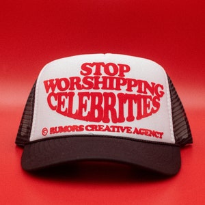 May include: A black and white trucker hat with the slogan "STOP WORSHIPPING CELEBRITIES" in red. The hat features a white front panel, a black peak, and a mesh back. The text is in a bold, stylized font.