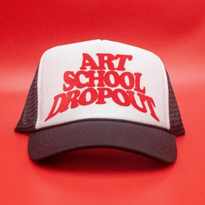 May include: A black and white trucker hat with the words "ART SCHOOL DROPOUT" in red block letters. The hat features a mesh back and a curved peak.