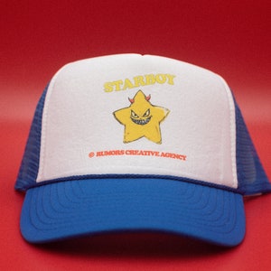 May include: A blue and white trucker hat featuring the word "STARBOY" in yellow, above a yellow star with a devilish face. The hat has a blue peak and mesh back. The text "© RUMORS CREATIVE AGENCY" is printed below the star.