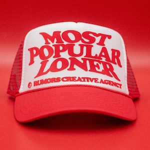 May include: A red and white trucker hat featuring the words "MOST POPULAR LONER" in red. The hat has a red peak and mesh back, with "RUMORS CREATIVE AGENCY" printed below the main text.