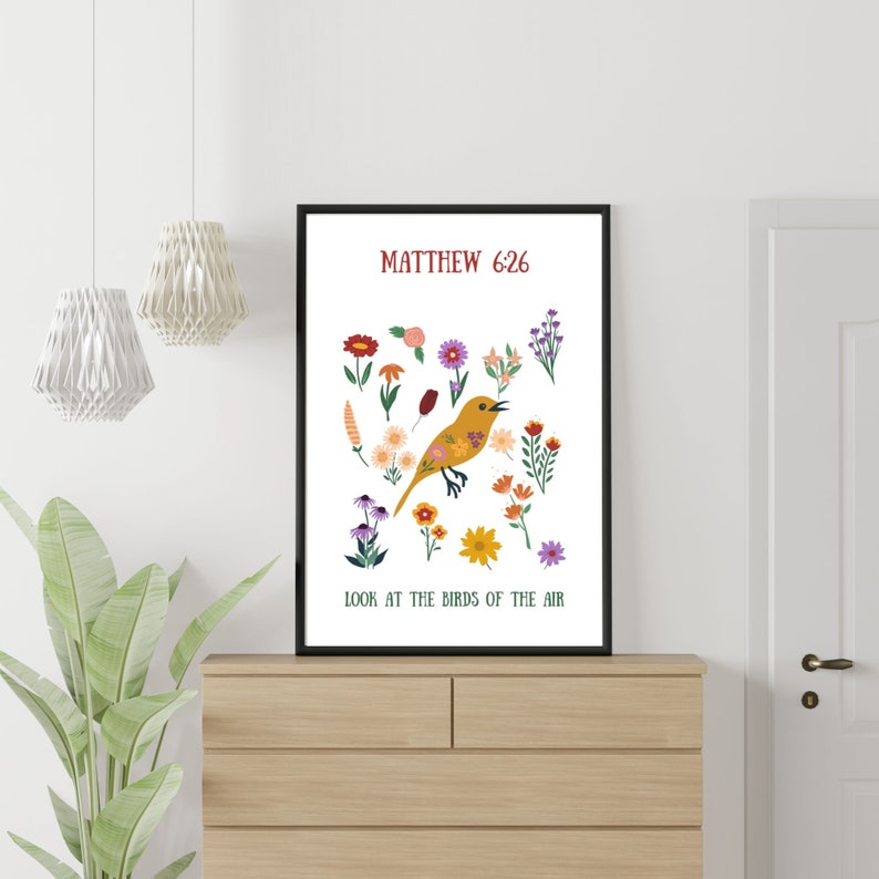 May include: A framed art print with a yellow bird illustration and floral elements. The text reads "Matthew 6:26" and "Look at the birds of the air". The print is displayed on a wooden chest of drawers. The print is in a black frame.