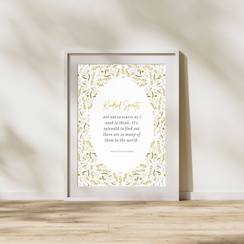 May include: A framed print with the quote "Kindred Spirits" in gold script, surrounded by a floral border of green and pink leaves. The quote is from Anne of Green Gables. The frame is white.