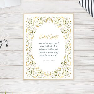 May include: A framed print featuring the quote "Kindred Spirits" from Anne of Green Gables. The text is in a gold script font, surrounded by a floral border with green leaves and pink flowers. The frame is a light beige color.