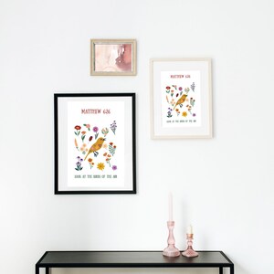 May include: Three framed art prints on a white wall. The prints feature a yellow bird illustration surrounded by colourful flowers, with the text "Matthew 6:26" and "Look at the birds of the air." A small framed watercolour and two pink candle holders are also visible.
