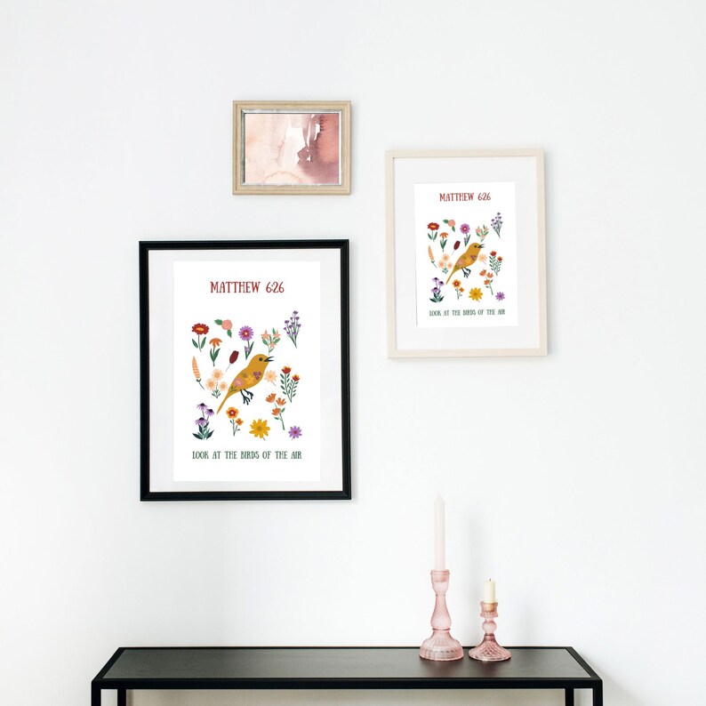 May include: Three framed art prints on a white wall. The prints feature a yellow bird illustration surrounded by colourful flowers, with the text "Matthew 6:26" and "Look at the birds of the air." A small framed watercolour and two pink candle holders are also visible.