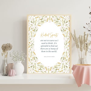 May include: A framed print with the quote "Kindred Spirits" in gold script, surrounded by a floral border of green leaves and pink flowers. The print is displayed on a white shelf with decorative vases and dried flowers.
