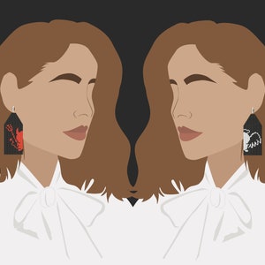 May include: A woman with brown hair is wearing a white shirt with a bow tie. She is wearing a pair of black earrings with red and white embroidered designs. The earrings are shaped like a rectangle.