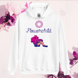 Op de afbeelding: Witte hoodie met een roze en paarse flowerchild-print. Het ontwerp toont een persoon die zit met een grote roos boven zijn hoofd. De tekst "Flowerchild" is in paars boven het ontwerp gedrukt.